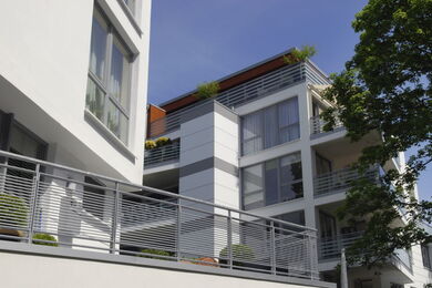 Moderner Wohnblock mit großen Fenstern, Balkonen und Metallgeländern, gesehen an einem sonnigen Tag mit blauem Himmel und einem belaubten Baum, der teilweise rechts zu sehen ist.
