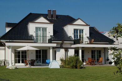 Ein modernes zweistöckiges Haus mit dunklen Dachziegeln, weißen Außenwänden, Balkonen und großen Fenstern. Es gibt einen grünen Rasen mit Gartenmöbeln, einen Sonnenschirm und ein kleines Zelt im hinteren Garten.
