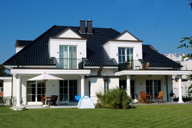 Ein modernes zweistöckiges Haus mit dunklem Ziegeldach, weißen Wänden, Balkonen und großen Fenstern in einem grünen, grasbewachsenen Garten mit Terrassenmöbeln, einem Sonnenschirm und einem kleinen Zelt. Der Himmel ist klar und blau.