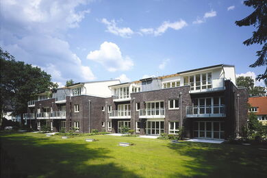 Ein modernes, dreistöckiges Wohnhaus mit großen Fenstern und Balkonen steht vor einer gepflegten Rasenfläche unter einem teilweise bewölkten Himmel. Bäume begrenzen das Grundstück.