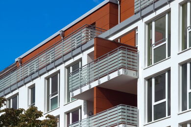 Moderner Wohnblock mit weißen Wänden, großen Fenstern und Balkonen mit Metallgeländern. Die Fassade weist braune Akzentplatten auf, und im Hintergrund ist der Himmel klar und blau.
