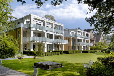 Modern three-storey block of flats with large balconies overlook a spacious, well-kept green lawn bordered by trees and shrubs, under a partly cloudy sky.