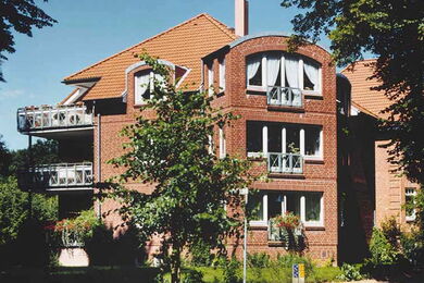 A three-storey brick block of flats with white-framed windows, small balconies, and a red-tiled roof, surrounded by greenery and trees on a sunny day.
