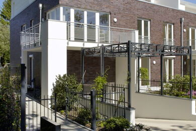 Modernes Stadthaus mit Backstein und weißem Äußeren, Glasbalkon, großen Fenstern, Metallzaun und einer Pergola über dem Eingang, umgeben von Grün und Sträuchern.