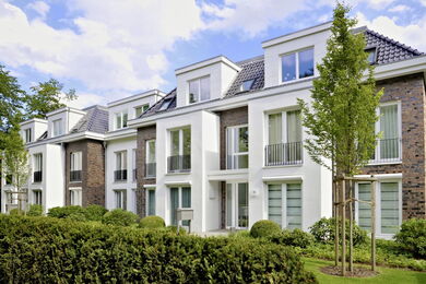Modern three-storey block of flats with large windows, small balconies, and a mix of white and dark brick facades, surrounded by greenery and neatly trimmed bushes under a partly cloudy sky.