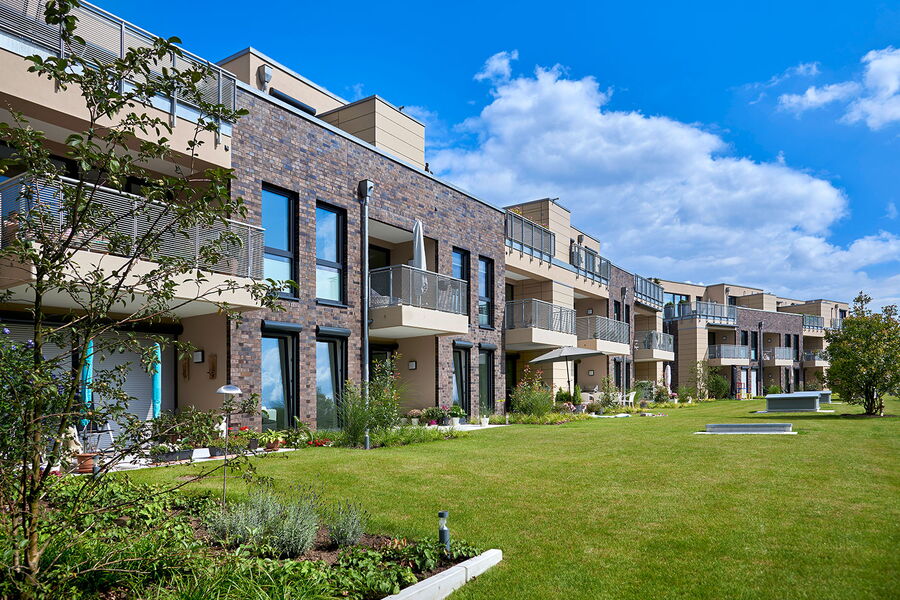 Moderner Wohnblock mit großen Balkonen und Ziegelsteinakzenten, umgeben von einem gepflegten grünen Rasen und angelegten Gärten unter einem strahlend blauen Himmel mit einigen Wolken.