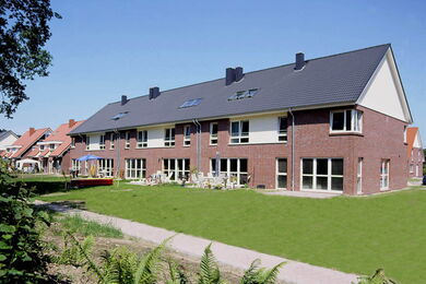Eine Reihe moderner Backstein-Reihenhäuser mit dunklen Dächern und großen Fenstern, die auf einen gepflegten grünen Rasen und einen kleinen Fußweg unter einem klaren blauen Himmel blicken.