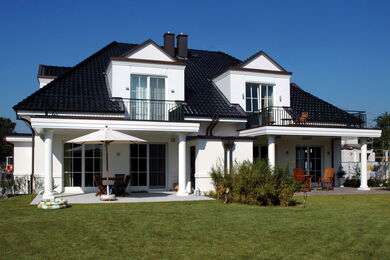 Modernes zweigeschossiges weißes Haus mit schwarzem Dach, großen Fenstern und Balkonen. Es gibt eine Terrasse mit Tisch, Stühlen, Sonnenschirm und Sitzgelegenheiten im Freien, umgeben von einem gepflegten Rasen und Sträuchern unter einem klaren blauen Him