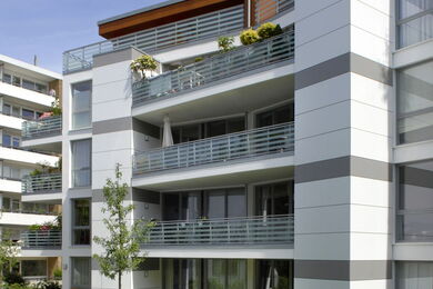 Modernes Mehrfamilienhaus mit mehreren Etagen und Balkonen, mit Glasgeländern, weißen und grauen Außenverkleidungen und einigen Pflanzen und Gartenmöbeln auf den Balkonen.