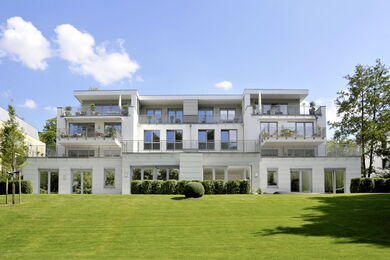 Modern white three-storey apartment building with large windows and balconies, set on a well-maintained, green lawn with a few small trees under a blue sky with scattered clouds.