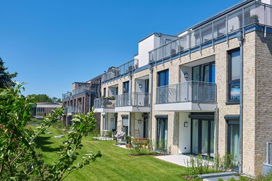Moderne Wohnblocks mit Balkonen und großen Fenstern, umgeben von grünen Rasenflächen und kleinen Sträuchern, unter einem klaren blauen Himmel.