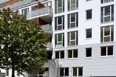 Ein moderner Wohnblock mit großen Fenstern und Balkonen, teilweise verdeckt durch einen grünen Baum im Vordergrund. Das Gebäude hat eine weiß-graue Fassade.