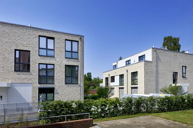 Two modern, light brick block of flats with large windows are separated by greenery and a bush-lined path, under a clear blue sky.