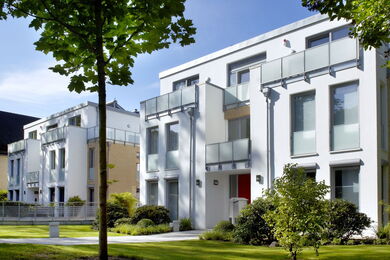 Modern white apartment blocks with large windows and glass balconies, surrounded by green lawns, trees, and shrubs on a sunny day.