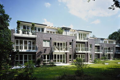 Ein moderner, dreistöckiger Wohnblock mit dunklen Backsteinmauern, großen Fenstern und mehreren Balkonen, die auf eine Wiese blicken, umgeben von Bäumen unter einem teilweise bewölkten Himmel.
