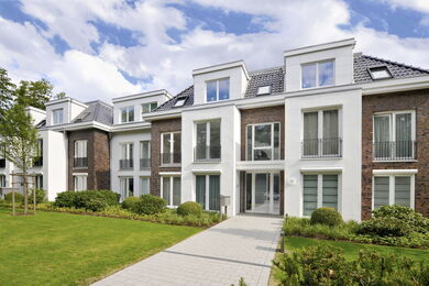 Modern, multi-storey residential building with large windows, brick and white exterior, and landscaped front garden with green grass, shrubs, and a paved path leading to the entrance.