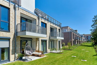 Moderner Wohnblock mit Balkonen, die an sonnigen Tagen einen Blick auf einen gepflegten grünen Rasen bieten. Im Erdgeschoss gibt es einen Hängesessel und Sitzgelegenheiten im Freien, und das Gebäude hat große Fenster und ein klares, modernes Design.
