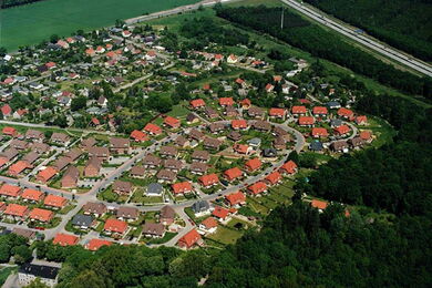 Aerial view of a suburban neighbourhood with rows of houses featuring red-tiled roofs, surrounded by trees and greenery. A road and open fields are visible in the background.