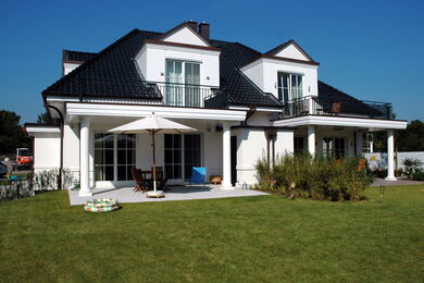 Ein modernes zweistöckiges weißes Haus mit dunklem Dach, zwei Balkonen, großen Fenstern und einem gepflegten grünen Rasen. Es gibt eine Veranda mit Gartenmöbeln und einem Sonnenschirm. Der Himmel ist klar und blau.