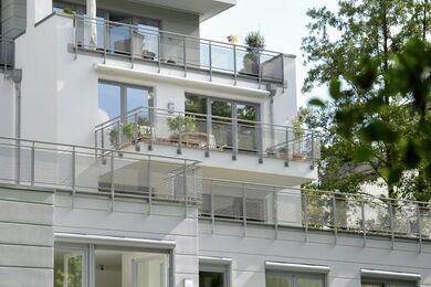 Modern apartment block with large windows and multiple balconies featuring potted plants and outdoor furniture. Trees and greenery are visible in the foreground. The building exterior is white and contemporary in design.