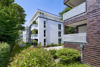 Modern apartment blocks with white and brown facades, large windows, and balconies surrounded by green bushes and trees on a sunny day.