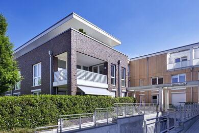 Modern two-storey building with brick and wood facades, large windows, balconies, and a covered entrance. A green hedge lines the front, and the sky is clear and blue.