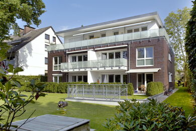 Modernes dreistöckiges Wohnhaus mit großen Balkonen und Glasgeländern, umgeben von grünem Rasen und Hecken, mit Bäumen und Nachbarhäusern im Hintergrund.