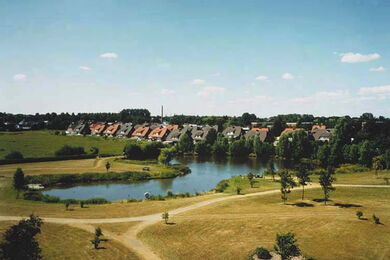 A small lake is surrounded by grassy paths and trees, with a row of houses featuring red roofs in the background under a blue sky.