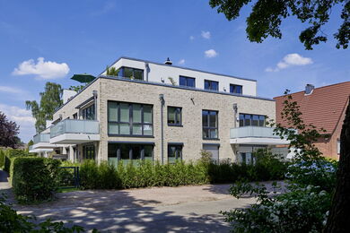 Modern three-storey block of flats with light brick exterior, large windows, and balconies, surrounded by greenery and trees, under a blue sky with scattered clouds.
