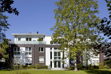 Ein moderner dreistöckiger Wohnblock mit großen Fenstern, einer Mischung aus dunklem und weißem Äußeren und einem gepflegten Rasen, der teilweise von einem hohen Baum beschattet wird, unter einem klaren blauen Himmel.