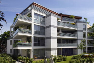 Moderner Wohnblock mit mehreren Balkonen, großen Fenstern und horizontalen grauen Akzenten. Umgeben von Grünflächen und einem gepflegten Rasen steht das Gebäude vor einem klaren blauen Himmel.