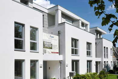 A modern white block of flats with large windows, flat roofs, and balconies. A banner hangs on the façade, and green shrubs line the front. The sky is clear and blue, and a leafy tree branch is visible on the right.