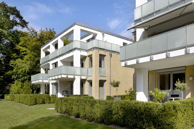 Modern apartment block with large glass balconies, white and beige exterior, surrounded by green hedges and a well-maintained lawn on a sunny day.