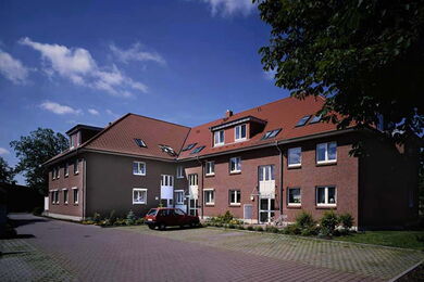 Ein dreistöckiges Wohnhaus aus Backstein mit einem roten Schrägdach, mehreren Fenstern und einem kleinen Parkplatz davor, auf dem ein rotes Auto geparkt ist. Der Himmel ist klar mit vereinzelten Wolken und im Hintergrund sind Bäume zu sehen.