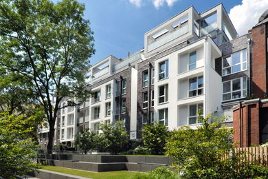 Modern apartment block with large windows and balconies, surrounded by trees, greenery, and landscaped gardens on a sunny day with a blue sky.