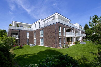 Modern three-storey block of flats with brick and white exterior, large balconies, many windows, and a well-maintained green lawn surrounded by trees and shrubs under a clear blue sky.