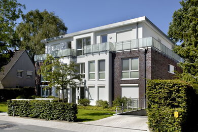 Modernes dreistöckiges Wohnhaus mit großen Fenstern, Glasbalkonen und einer Mischung aus weißem und dunklem Backstein, umgeben von Grünflächen und Hecken an einer ruhigen Straße.