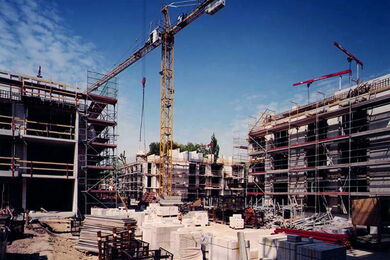 Baustelle mit Kränen, Gerüsten und teilweise gebauten mehrstöckigen Gebäuden unter blauem Himmel. Baumaterialien und Geräte sind auf der Baustelle verstreut.