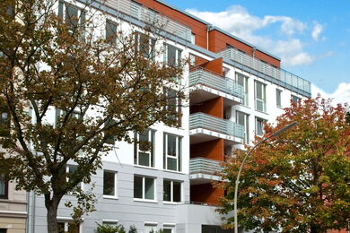 Modernes, mehrstöckiges Wohnhaus mit weißer, holzgetäfelter Fassade, mehreren Balkonen und großen Fenstern, die teilweise von Bäumen unter einem blauen Himmel mit Wolken verdeckt werden.