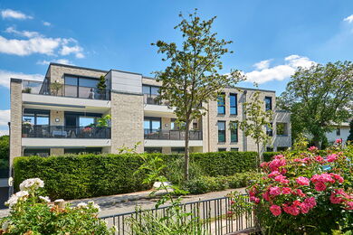Modernes dreistöckiges Wohnhaus mit Balkonen, umgeben von grünen Hecken und Bäumen. Bunte Blumen und Sträucher stehen im Vordergrund unter einem hellen, sonnigen Himmel mit ein paar weißen Wolken.