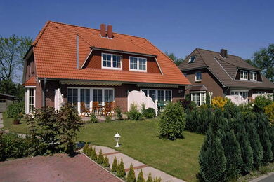 Two modern suburban houses with steeply sloped roofs, front patios, and well-kept gardens, set against a clear blue sky and surrounded by green lawns and shrubs.