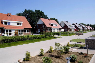 Eine Reihe moderner, zweistöckiger Häuser mit roten und weißen Dächern säumen eine ruhige Vorstadtstraße, gesäumt von grünem Rasen, kleinen Sträuchern und einem klaren blauen Himmel über uns.