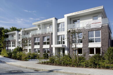 Modern three-storey block of flats with large windows, balconies, and a mix of white and dark brick exterior. Small trees and shrubs line the front, and a pavement runs along the street under a clear sky.