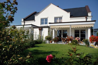 Ein modernes weißes Haus mit einem dunklen Dach, großen Fenstern und einer überdachten Terrasse, umgeben von einem üppigen grünen Rasen und bunten Blumen unter einem klaren blauen Himmel. Ein Tisch und Stühle sind auf der Terrasse zu sehen.