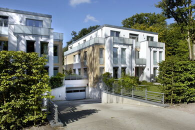 A modern, white three-storey block of flats with large windows, balconies with glass balustrades, and a red front door. The building is surrounded by greenery and has a paved path leading to the entrance.