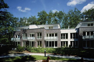 Modernes zweistöckiges Wohnhaus mit Balkonen, großen Fenstern und umgeben von Bäumen unter blauem Himmel mit vereinzelten Wolken.