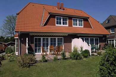 Two-storey brick house with a red tiled roof, dormer windows, and a divided patio area with outdoor furniture. The house is surrounded by a green lawn and some bushes, under a clear blue sky.