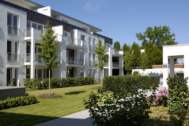 Moderne weiße Wohnblöcke mit Balkonen umgeben einen begrünten Innenhof mit Rasen, kleinen Bäumen, Hecken und Sträuchern unter einem klaren blauen Himmel.