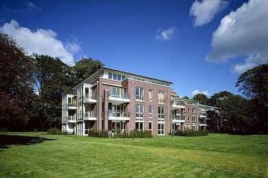 Ein moderner dreistöckiger Wohnblock mit großen Fenstern und Balkonen, umgeben von einem gepflegten grünen Rasen und Bäumen, unter einem strahlend blauen Himmel mit vereinzelten Wolken.
