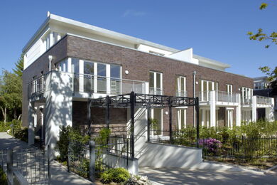 Ein modernes Stadthaus mit weißer Backsteinfassade, großen Fenstern, Balkonen mit Glasgeländern und einer Metallpergola über dem Eingang. Das Gebäude ist von Grün umgeben und steht vor einem klaren blauen Himmel.